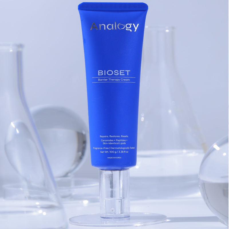 A blue tube of Analogy Bioset Sensory Therapy Cream stands upright on a clear acrylic stand, with blurred glass laboratory equipment in the background.